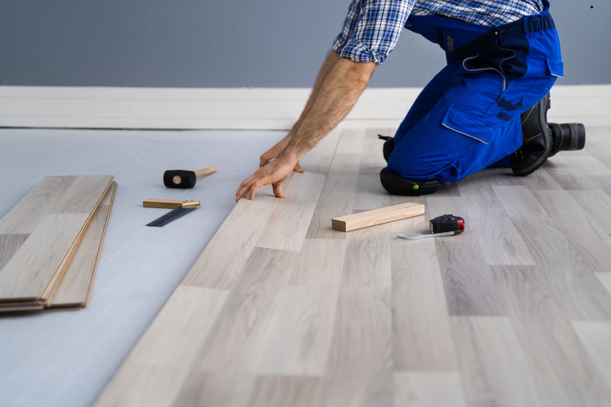 A person in blue overalls installs wooden flooring, using various tools like a mallet, measuring tape, and wooden spacers.