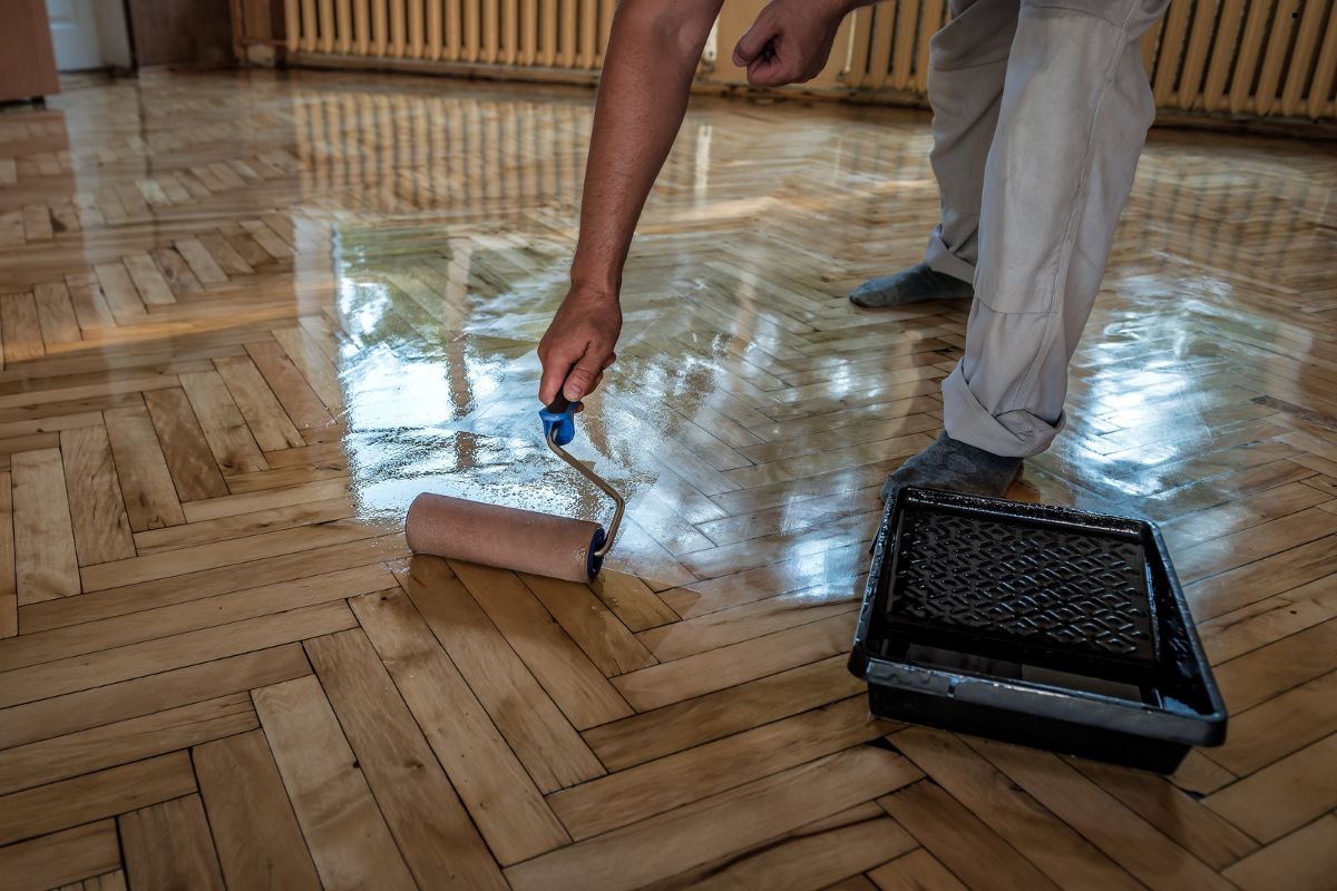 A person applies varnish to a polished wooden floor with a paint roller, showcasing careful renovation in a well-lit room.