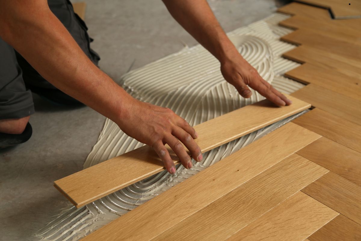 A worker carefully lays a wooden floorboard onto adhesive, blending craftsmanship and technique for a seamless finish.